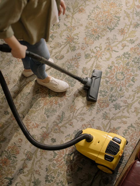 A person using a yellow vacuum cleaner with a black hose to perform surface cleaning on an ornate, patterned rug in a living room or similar indoor space. The rug features floral motifs in muted shades of beige, green, and orange, laid on a hard flooring base. The individual, dressed in casual clothing and white sneakers, is slightly bent over, guiding the vacuum cleaner across the textured surface. Bright indoor lighting highlights the cleanliness and detail of the rug. The setting appears well-maintained, consistent with professional domestic cleaning services provided by Highbury Carpet Cleaners, as referenced in the N5 carpet cleaning guide for Highbury Fields homes. The image emphasizes deep cleaning and sanitisation processes crucial for maintaining hygienic and visually appealing surfaces within residential spaces.